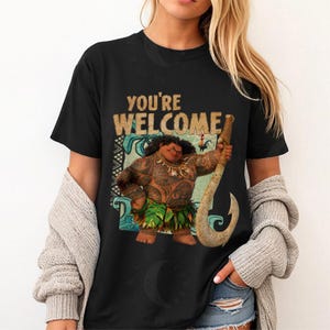 Peut inclure: T-shirt noir avec une illustration de dessin anim&eacute; d'un personnage polyn&eacute;sien tenant un grand crochet. Le texte "YOU'RE WELCOME" est imprim&eacute; au-dessus du personnage. Le personnage est brun avec des tatouages et une jupe en herbe.