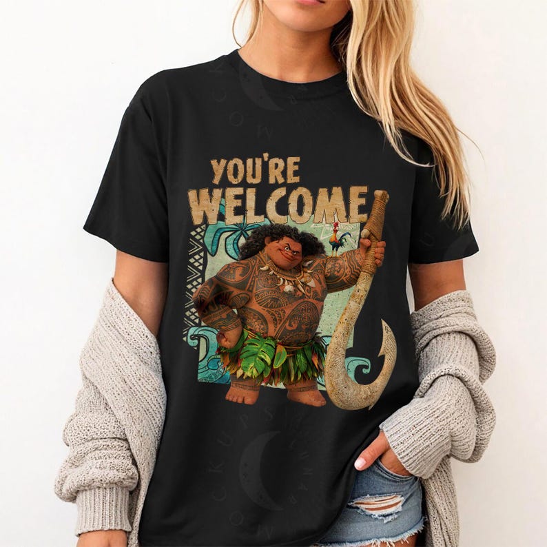 Peut inclure: T-shirt noir avec une illustration de dessin anim&eacute; d'un personnage polyn&eacute;sien tenant un grand crochet. Le texte "YOU'RE WELCOME" est imprim&eacute; au-dessus du personnage. Le personnage est brun avec des tatouages et une jupe en herbe.