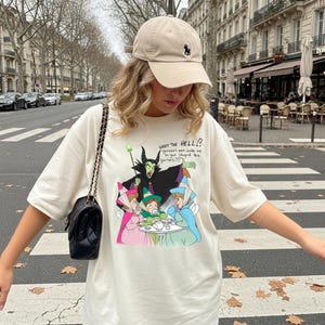 Puede incluir: Camiseta color crema con un gr&aacute;fico de dibujos animados de un villano con un bast&oacute;n verde y tres figuras en una mesa. El texto "WHAT THE HELL!?" y otro texto est&aacute; encima del gr&aacute;fico. Se usa una gorra de b&eacute;isbol beige.