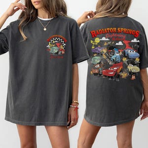 Pode incluir: T-shirt cinza escuro com um gr&aacute;fico colorido de personagens do filme Carros. A frente da camisa tem o texto "Radiator Springs" e o nome "Jessica". A parte de tr&aacute;s da camisa tem um gr&aacute;fico maior com "Radiator Springs Lightning McQueen".