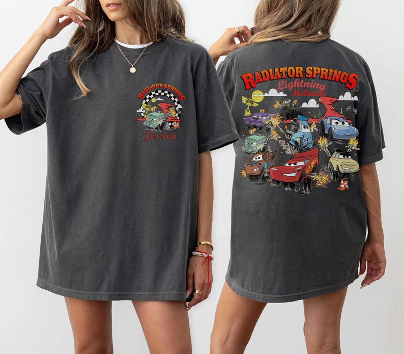 Pode incluir: T-shirt cinza escuro com um gr&aacute;fico colorido de personagens do filme Carros. A frente da camisa tem o texto "Radiator Springs" e o nome "Jessica". A parte de tr&aacute;s da camisa tem um gr&aacute;fico maior com "Radiator Springs Lightning McQueen".