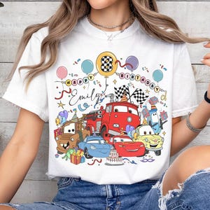 May include: White t-shirt featuring a colorful graphic of cartoon cars, balloons, and the text "Happy Birthday Emily." The design includes a birthday cake, checkered flags, and gift boxes, creating a festive theme.