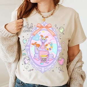 May include: Beige t-shirt featuring a cartoon dragon inside a decorative frame. The dragon holds a palette and paint bucket. The text "one little spark" is above the frame, and "Figment Pigment" is on the bucket.