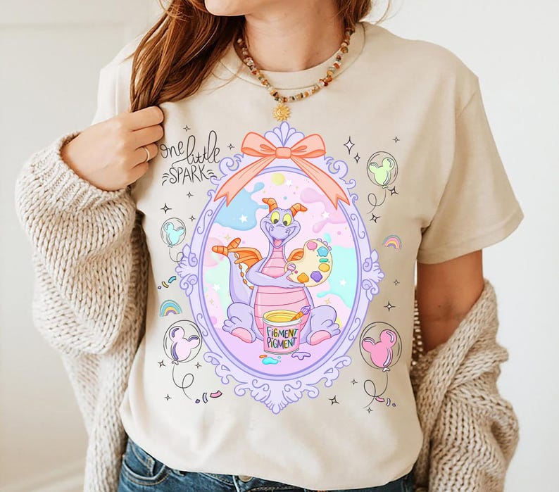 May include: Beige t-shirt featuring a cartoon dragon inside a decorative frame. The dragon holds a palette and paint bucket. The text "one little spark" is above the frame, and "Figment Pigment" is on the bucket.