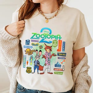 Op de afbeelding: Beige T-shirt met het logo van de Disney-film "Zootopia 2" en de personages Judy Hopps en Nick Wilde. Het shirt toont kleurrijke afbeeldingen van locaties uit de film, zoals Tundratown en Sahara Square.