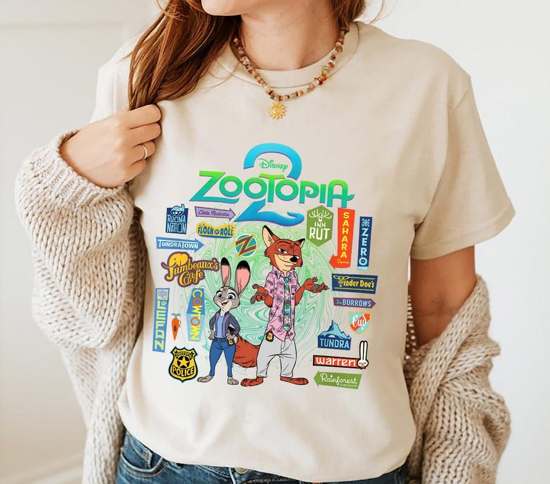 Op de afbeelding: Beige T-shirt met het logo van de Disney-film "Zootopia 2" en de personages Judy Hopps en Nick Wilde. Het shirt toont kleurrijke afbeeldingen van locaties uit de film, zoals Tundratown en Sahara Square.