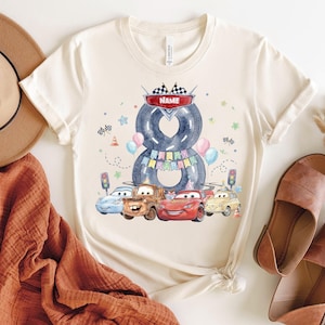 May include: Cream-colored t-shirt featuring a colorful graphic design. The design includes the number 8 formed by two tires, a banner that says "Name", and cartoon cars. The shirt is decorated with balloons, stars, and traffic lights.