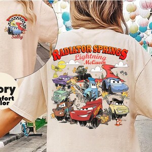 Pode incluir: Camiseta de cor de conforto marfim com um gr&aacute;fico colorido de personagens do filme Carros, incluindo Rel&acirc;mpago McQueen e Mate, com o texto "Radiator Springs Lightning McQueen". Um gr&aacute;fico menor est&aacute; na parte superior das costas.