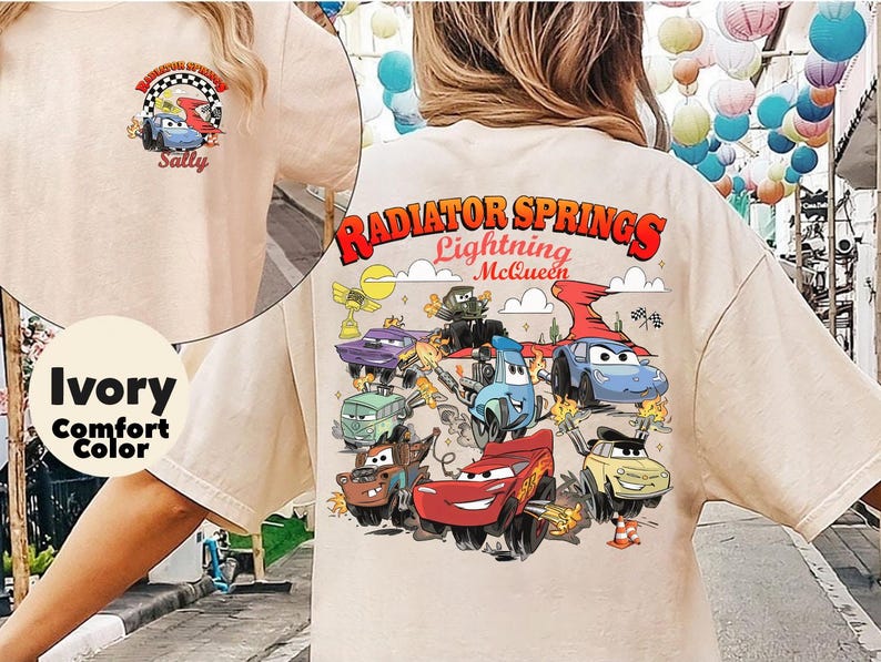Pode incluir: Camiseta de cor de conforto marfim com um gr&aacute;fico colorido de personagens do filme Carros, incluindo Rel&acirc;mpago McQueen e Mate, com o texto "Radiator Springs Lightning McQueen". Um gr&aacute;fico menor est&aacute; na parte superior das costas.
