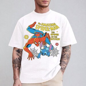 May include: White t-shirt featuring a colorful graphic of Spider-Man and Stitch. The design includes the text "THE AMAZING SPIDER-MAN THE SPIDER OR THE STITCH?" with comic book style elements. The shirt has short sleeves and a crew neck.