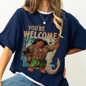 Peut inclure: T-shirt bleu marine avec l'inscription "YOU'RE WELCOME" et un dessin d'un personnage polyn&eacute;sien avec des tatouages et une jupe en herbe, tenant un crochet. V&ecirc;tement d&eacute;contract&eacute;.