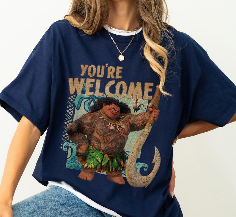 Peut inclure: T-shirt bleu marine avec l'inscription "YOU'RE WELCOME" et un dessin d'un personnage polyn&eacute;sien avec des tatouages et une jupe en herbe, tenant un crochet. V&ecirc;tement d&eacute;contract&eacute;.