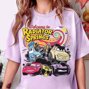 Może przedstawiać: Jasnofioletowa koszulka z napisem "Welcome to Radiator Springs" w kolorze ż&oacute;łtym i pomarańczowym. Grafika przedstawia kresk&oacute;wkowe samochody w r&oacute;żnych kolorach na tle kolorowych wir&oacute;w.