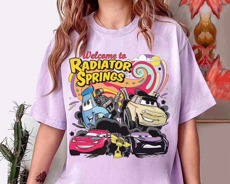 Może przedstawiać: Jasnofioletowa koszulka z napisem "Welcome to Radiator Springs" w kolorze ż&oacute;łtym i pomarańczowym. Grafika przedstawia kresk&oacute;wkowe samochody w r&oacute;żnych kolorach na tle kolorowych wir&oacute;w.