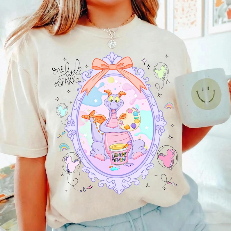 May include: Cream-colored t-shirt featuring a whimsical illustration of a purple dragon holding a palette and paint bucket. The design is framed within a decorative oval, with the words "one little spark" above. A white mug with a smiley face is also visible.
