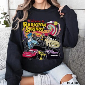 Może przedstawiać: Czarna bluza z kolorową grafiką samochod&oacute;w z kresk&oacute;wek i napisem "Welcome to Radiator Springs". Projekt zawiera niebieski samoch&oacute;d, beżowy samoch&oacute;d, czerwony samoch&oacute;d i fioletowy samoch&oacute;d. Bluza ma okrągły dekolt.