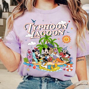 May include: A light purple t-shirt featuring the words "Typhoon Lagoon" in a retro font, with cartoon characters on a beach scene. The design includes palm trees, a sun, and characters like Mickey and Minnie Mouse.