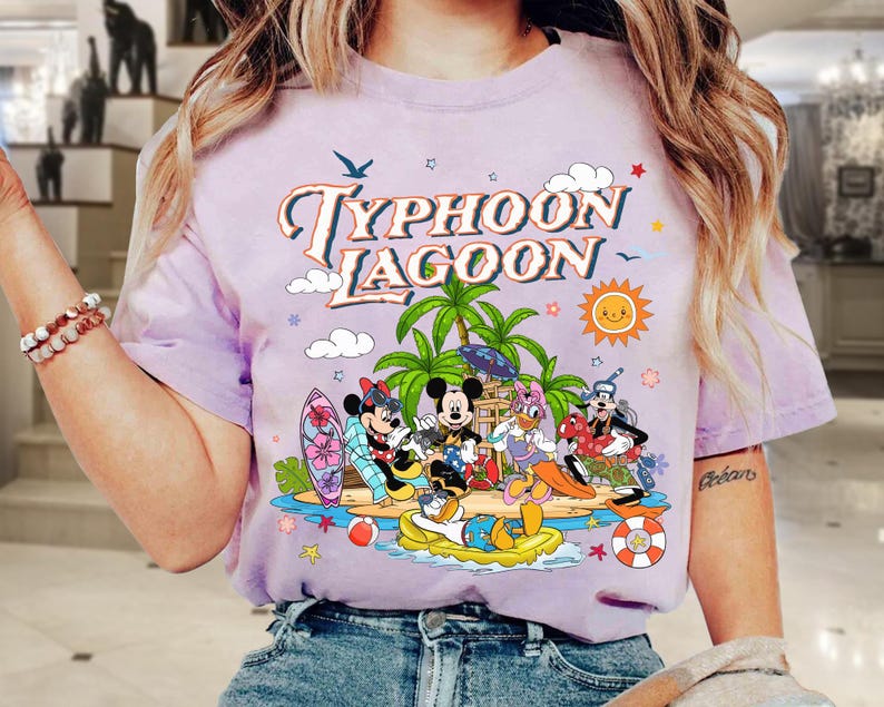 May include: A light purple t-shirt featuring the words "Typhoon Lagoon" in a retro font, with cartoon characters on a beach scene. The design includes palm trees, a sun, and characters like Mickey and Minnie Mouse.