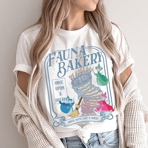 May include: White t-shirt featuring a whimsical design for "Fauna Bakery." The design includes a multi-layered cake with candles, and three fairy characters. The text reads "Once Upon a Dream" and "Baked with Love & Magic Since 1959."