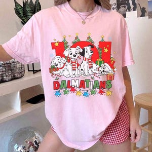 May include: A light pink short-sleeve t-shirt featuring a graphic of four Dalmatian puppies wearing red and white scarves and Santa hats. The design includes the text 101 DALMATIANS, Christmas trees, wrapped gifts, and colorful holiday lights.