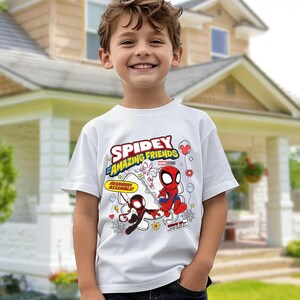 Puede incluir: Una camiseta blanca con el texto "Spidey and His Amazing Friends" e ilustraciones de dibujos animados de Spider-Man y otros personajes. La camiseta tiene mangas cortas y la lleva un ni&ntilde;o.