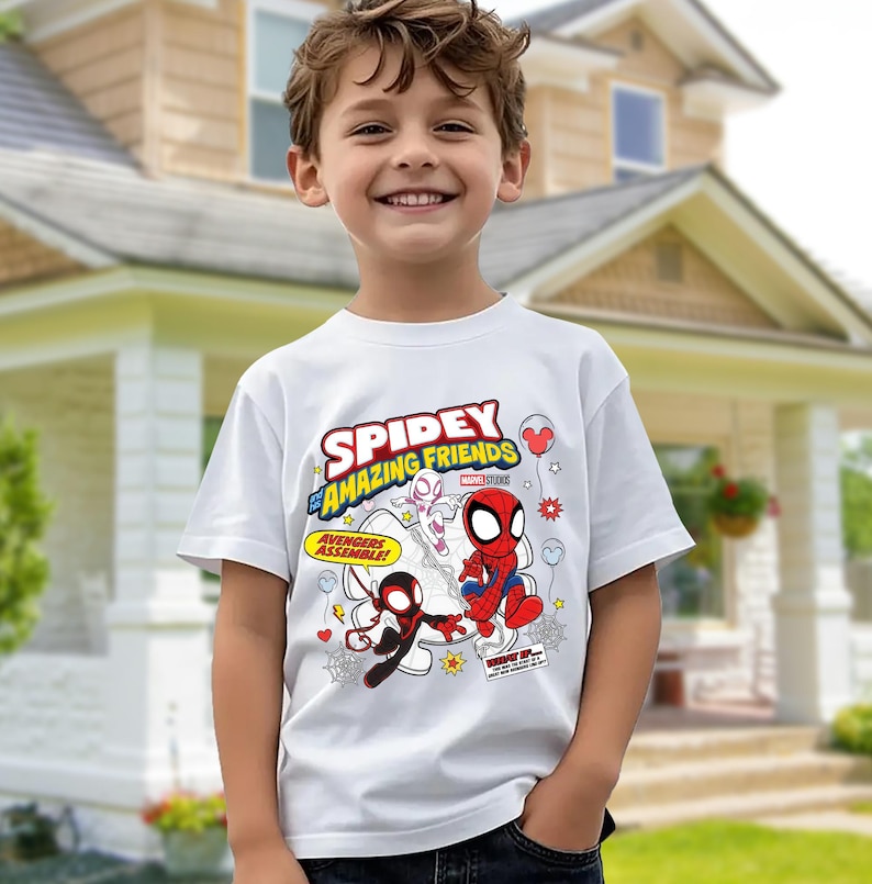 Puede incluir: Una camiseta blanca con el texto "Spidey and His Amazing Friends" e ilustraciones de dibujos animados de Spider-Man y otros personajes. La camiseta tiene mangas cortas y la lleva un ni&ntilde;o.
