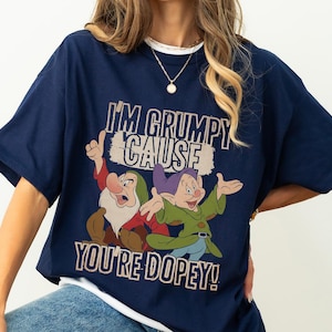 May include: Navy blue t-shirt with a cartoon graphic of two dwarfs and the text "I'M GRUMPY CAUSE YOU'RE DOPEY!". The shirt has a white trim. The graphic features red, green, purple, and brown colours. The t-shirt is a casual wear item.