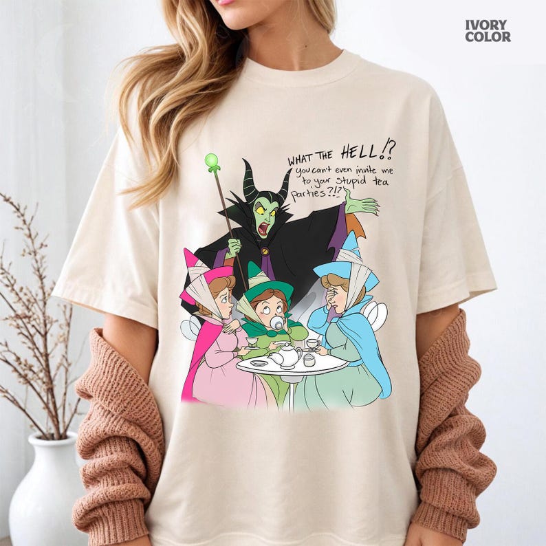Puede incluir: Camiseta de color marfil con una ilustraci&oacute;n de dibujos animados de un villano y tres hadas en una fiesta de t&eacute;. El villano tiene la piel verde con cuernos negros y una capa negra. El texto de la camiseta dice "WHAT THE HELL!?" y "you can't even invite me to your stupid tea parties?!?".