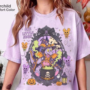 Puede incluir: Una camiseta color orquídea con un gráfico de un dragón morado con sombrero de brujo y capa, revolviendo un caldero negro con diseño de calavera. El texto dice One Little Spark, rodeado de globos de calabaza y un marco oscuro ornamentado.
