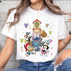 May include: White t-shirt featuring a colorful cartoon graphic with characters from Looney Tunes and Disney. The design includes a water tower with the WB logo, Stitch, and other animated figures. The shirt has a crew neck.
