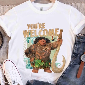 Peut inclure: T-shirt blanc avec un personnage de dessin anim&eacute; tenant un grand crochet. Le texte "YOU'RE WELCOME" est imprim&eacute; au-dessus du personnage. Le personnage a des tatouages et porte une jupe en feuilles.