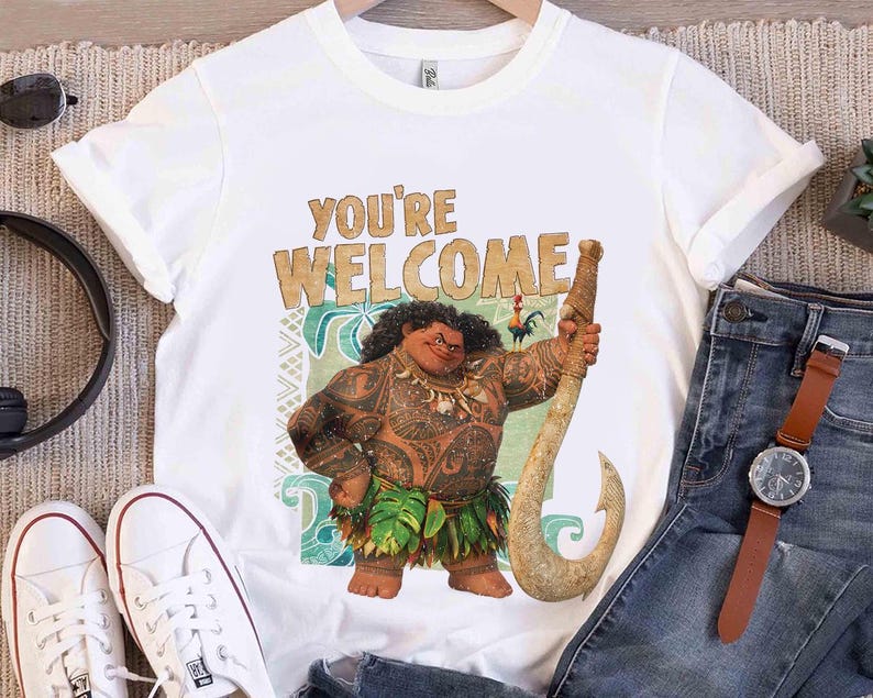 Peut inclure: T-shirt blanc avec un personnage de dessin anim&eacute; tenant un grand crochet. Le texte "YOU'RE WELCOME" est imprim&eacute; au-dessus du personnage. Le personnage a des tatouages et porte une jupe en feuilles.