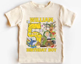 May include: Cream-colored short-sleeved t-shirt with a colorful graphic design. The design features the name "William", the number "5", and the words "Birthday Boy" in yellow. Cartoon characters and a birthday cake are also included.