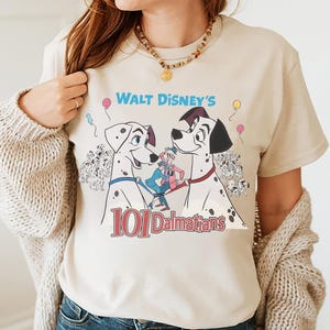 May include: Beige t-shirt featuring a graphic from Walt Disney's 101 Dalmatians. The design includes two Dalmatian dogs, the film's title, and other characters. The shirt has short sleeves and a crew neck.