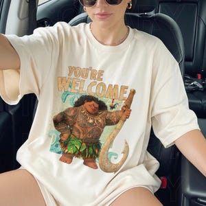 Peut inclure: T-shirt cr&egrave;me avec l'inscription "YOU'RE WELCOME" en beige. Le t-shirt pr&eacute;sente une illustration de dessin anim&eacute; d'un personnage tenant un grand crochet. Le personnage est brun avec des tatouages et une jupe verte.