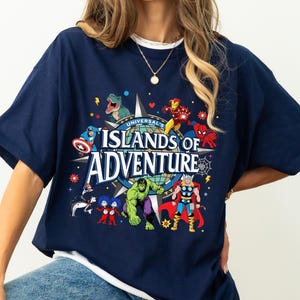 May include: Navy blue t-shirt featuring a colorful graphic of various Marvel characters and the text "Universal's Islands of Adventure." The design includes Iron Man, Spider-Man, Hulk, and Thor. The shirt has short sleeves and a crew neck.
