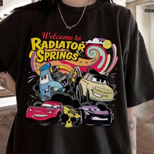 Może przedstawiać: Czarny t-shirt z kolorową grafiką z kresk&oacute;wek przedstawiającą samochody i napisem "Welcome to Radiator Springs". Projekt zawiera niebieski samoch&oacute;d, ż&oacute;łty samoch&oacute;d, czerwony samoch&oacute;d i fioletowy samoch&oacute;d na tle wirującej tęczy.