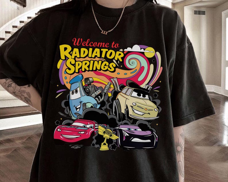 Może przedstawiać: Czarny t-shirt z kolorową grafiką z kresk&oacute;wek przedstawiającą samochody i napisem "Welcome to Radiator Springs". Projekt zawiera niebieski samoch&oacute;d, ż&oacute;łty samoch&oacute;d, czerwony samoch&oacute;d i fioletowy samoch&oacute;d na tle wirującej tęczy.