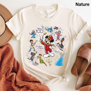 May include: Cream-colored t-shirt featuring Mickey Mouse in a red band uniform, conducting a musical ensemble of Disney characters. The shirt celebrates Disney's 100th anniversary with the text "100 Years of Music and Wonder."