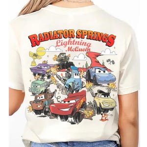 Pode incluir: Camiseta creme com um gr&aacute;fico colorido de personagens do filme Carros. O design inclui o texto "Radiator Springs Lightning McQueen" em vermelho e laranja, com v&aacute;rios personagens de carros em a&ccedil;&atilde;o.