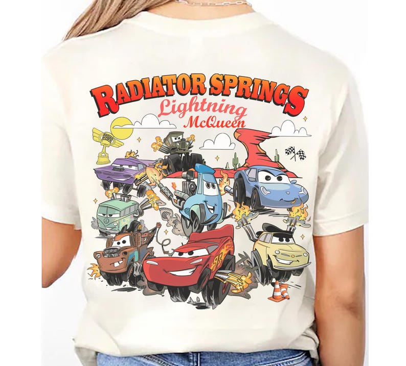 Pode incluir: Camiseta creme com um gr&aacute;fico colorido de personagens do filme Carros. O design inclui o texto "Radiator Springs Lightning McQueen" em vermelho e laranja, com v&aacute;rios personagens de carros em a&ccedil;&atilde;o.