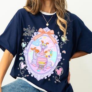 May include: Navy blue t-shirt featuring a whimsical illustration of a purple dragon holding a paint palette and a bucket labeled "Pigment Pigment." The design is framed within a decorative oval with a pink bow. The text "one little spark" is above the frame.