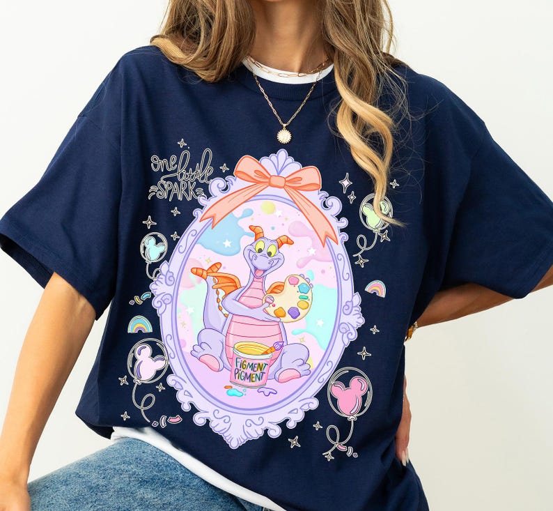 May include: Navy blue t-shirt featuring a whimsical illustration of a purple dragon holding a paint palette and a bucket labeled "Pigment Pigment." The design is framed within a decorative oval with a pink bow. The text "one little spark" is above the frame.
