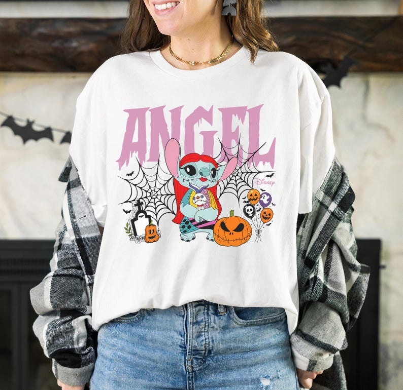 Pode incluir: Camiseta branca com um gr&aacute;fico de Halloween. O design inclui a palavra "ANGEL" em rosa, um personagem de desenho animado, teias de aranha, uma ab&oacute;bora esculpida e outros elementos de Halloween. A camiseta faz parte de uma cole&ccedil;&atilde;o da Disney.
