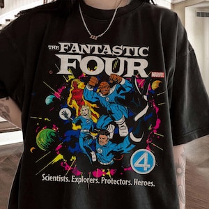 May include: Black t-shirt featuring a colorful graphic of The Fantastic Four comic book characters. The design includes the text "THE FANTASTIC FOUR" and "MARVEL", with the tagline "Scientists. Explorers. Protectors. Heroes."