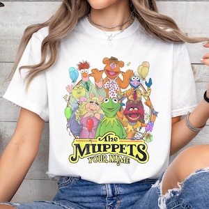 May include: White t-shirt featuring a colorful graphic of The Muppets characters, including Kermit the Frog, Miss Piggy, and Fozzie Bear. The shirt has the text "The Muppets" and "Your Name" in a yellow and black font.