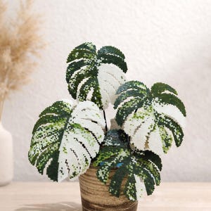 May include: A beaded Monstera plant in a brown ceramic pot. The leaves are a mix of white and green beads, creating a variegated pattern. The pot has a textured, earthy appearance, and the plant is set against a neutral background.