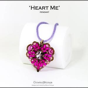 Può includere: Un ciondolo a forma di cuore con un cristallo trasparente centrale e perline rosa e marroni. Pende da un cordino lilla. Il testo "HEART ME" e "PENDANT" sono in alto.