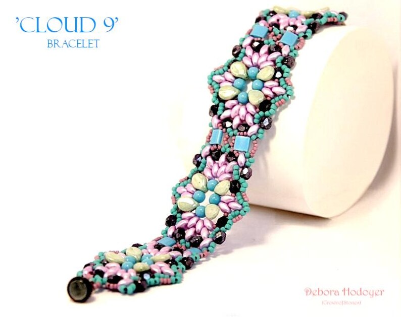 May include: A beaded bracelet with a floral design. The bracelet features a combination of pink, blue, and black beads. The text "CLOUD 9" and "BRACELET" are in the upper left corner.