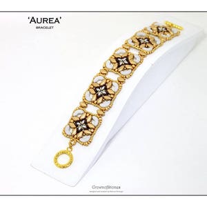May include: A gold and white beaded bracelet with the text "'AUREA' BRACELET" is displayed on a white stand. The bracelet features a floral design with gold and white beads. The clasp is gold, and the bracelet is designed by Crownofstones.